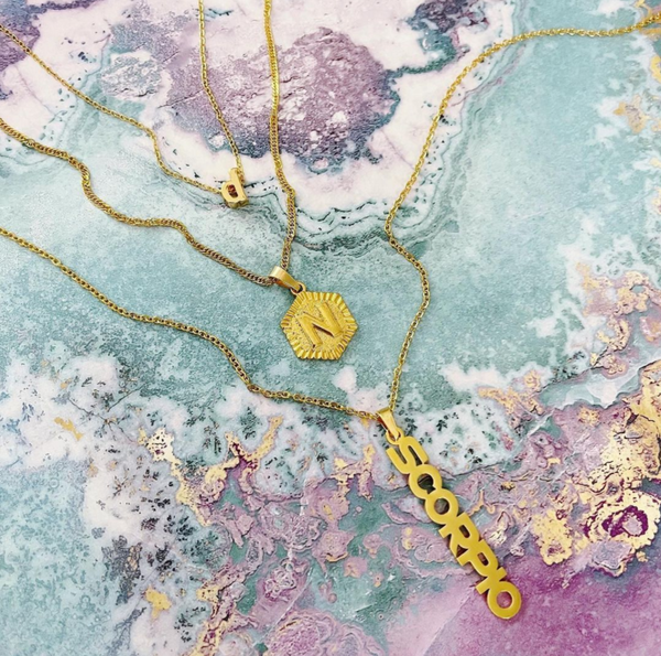 This is an image of our scorpio zodiac nameplate necklace layered with our mini letter and hexagon medallion necklaces on a blue and pink backgroun