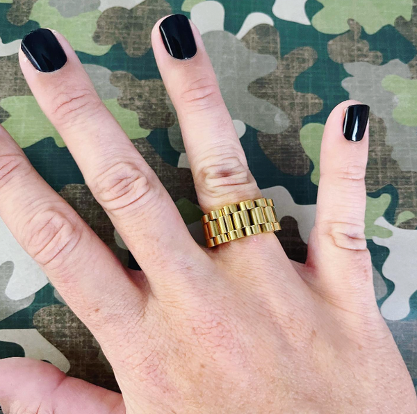 This is an image of a hand wearing our gold watchband style ring on a camoflauge background